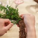 先以鐵絲穿過藤圈並收好，抓一把雪松，繞過後拉緊，再取一把雪松，在第一把根部接上，以此將雪松綁完。